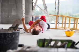 Slip and fall accident of a male worker at a construction site. An injured man lays on the floor, clutching his shoulder with a tipped over ladder next to him.
