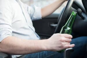 focus is on a man's arm while he is holding a beer bottle. The other arm is gripping a steering wheel inside a vehicle.