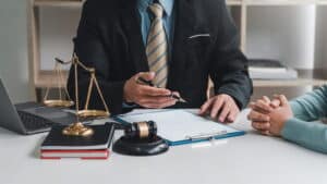 Closeup of a lawyer speaking with their client after a semi truck accident. He has a pen held over a clipboard with papers, and there's also a laptop, books, the scales of justice and a gavel on his desk.