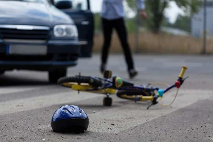 focus is on a bicycle and bicycle helmet laying on the ground after a car accident. A portion of the car is in the background with the driver standing next to it.