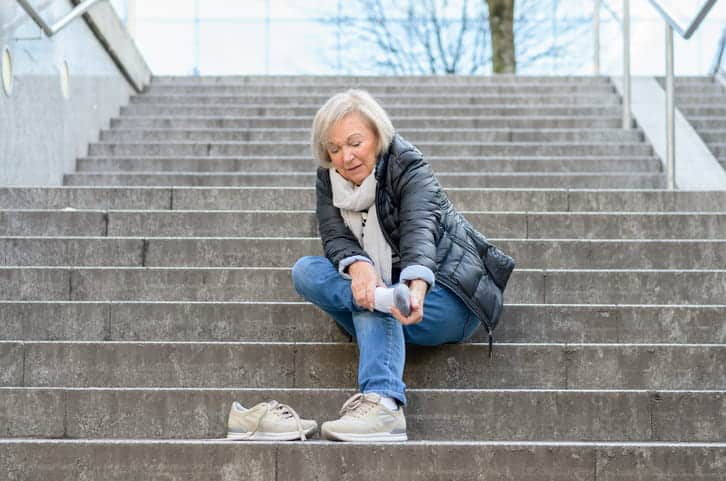 Focus is on an older woman who is sitting on the stairs with her shoe off, rubbing her ankle injury after she slipped and fell on the stairs.