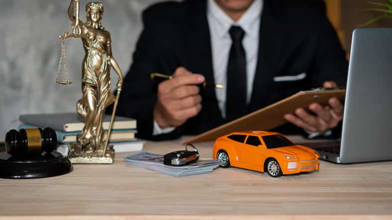 The focus is on a car accident lawyer, working on a case after a client hit a parked car. They're at their desk with paperwork, in front of a laptop. On their desk is a gavel, a statue of Lady Justice, car keys on a stack of money, books, and a toy car.