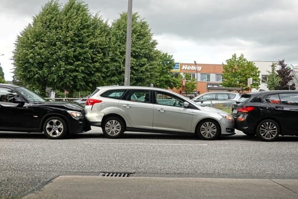 A Silver Station Wagon With Its Front End Pushed Into The Rear Of A Black Suv Ahead, While Its Own Rear Is Simultaneously Struck By Another Black Vehicle, Forming A Three-Car Chain Reaction Rear-End Collision On A City Street.