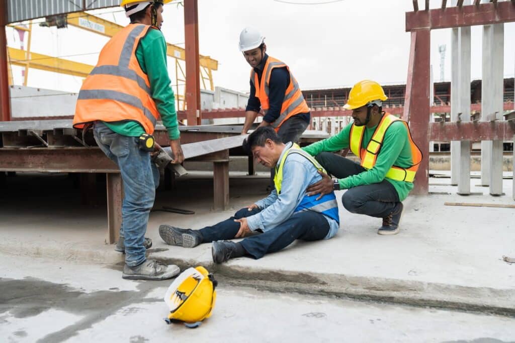 A construction worker sits on the ground after being hurt at work. Three other workers are around him ensuring he's okay.