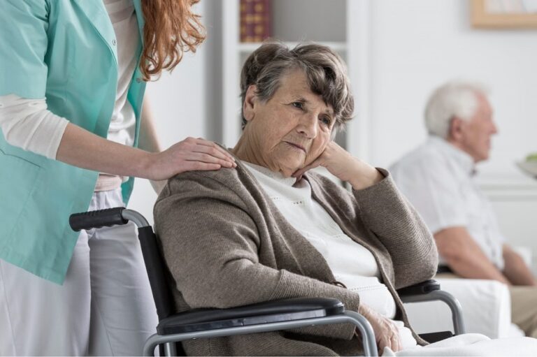 An Elderly Woman Sad In Her Wheelchair After Suffering From Nursing Home Abuse. An Aid Has Her Hand Resting On Her Shoulder.