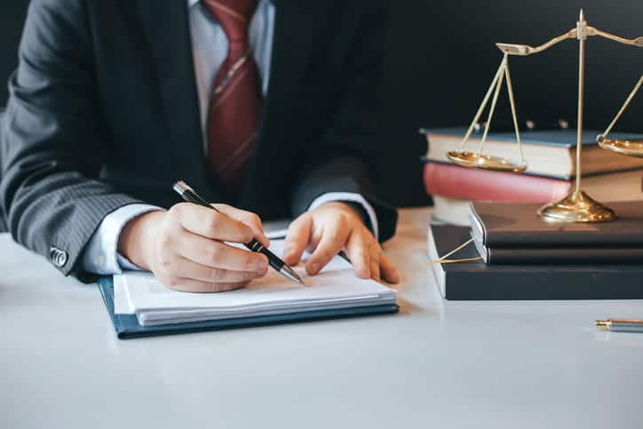 A Boat Injury Attorney Working On Paperwork At This Desk. Next To Him Is A Stack Of Books And The Scales Of Justice. 