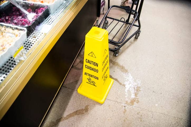 A slippery floor sign in a grocery store over a spill with a shopping car and customer nearby