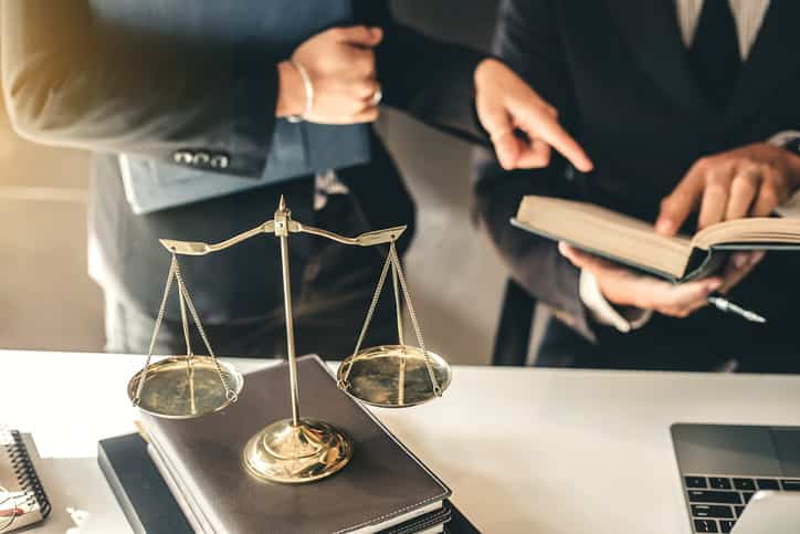 Two personal injury attorneys discuss how much a case is worth by reviewing a law textbook. On the desk are more books with the scales of justice. 