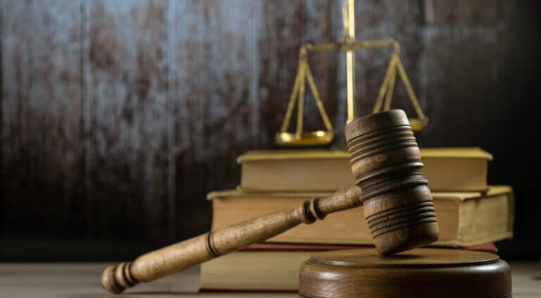 A Wooden Gavel In Front Of A Stack Of Law Books And The Scales Of Justice.