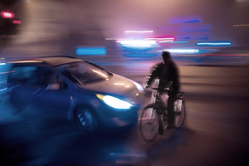A Blurred Image Showing A Nighttime Collision Between A Car And A Bicycle. The Scene Is Illuminated By Streetlights And The Headlights Of The Car, With Both Vehicles In Motion.
