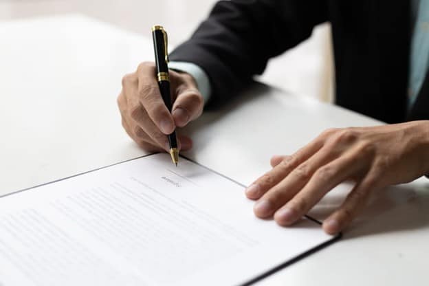 A Person In A Suit Signing A Document With A Pen, Representing The Process Of Filing A Police Report On A White Desk.