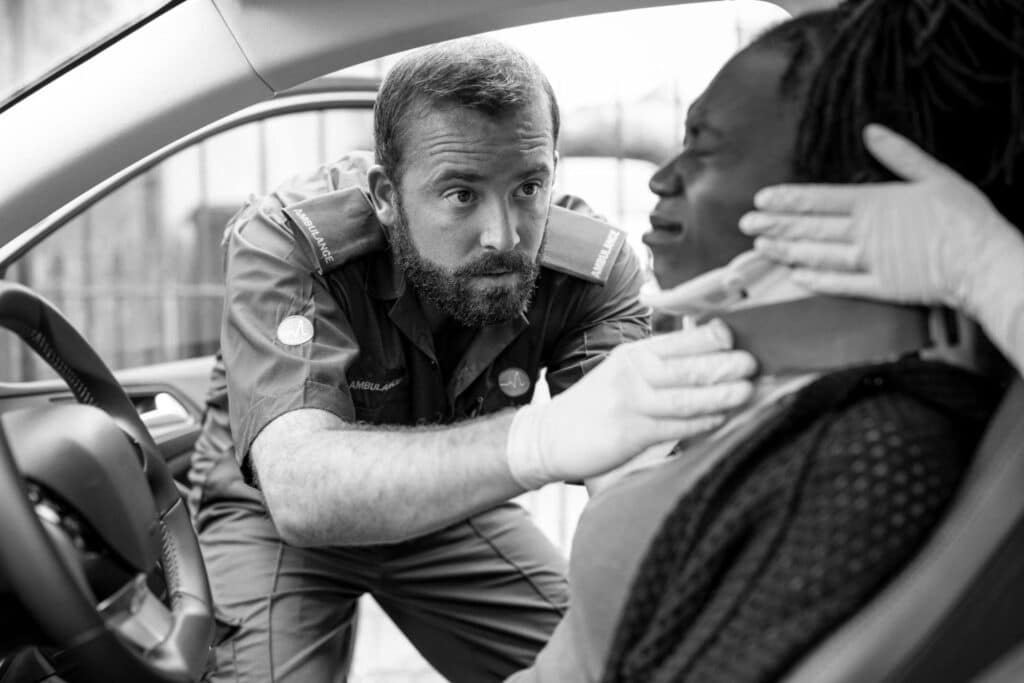 An Ambulance Crew Member In Uniform Treating A Patient Inside A Car, With Another Crew Member Assisting, In Black And White.