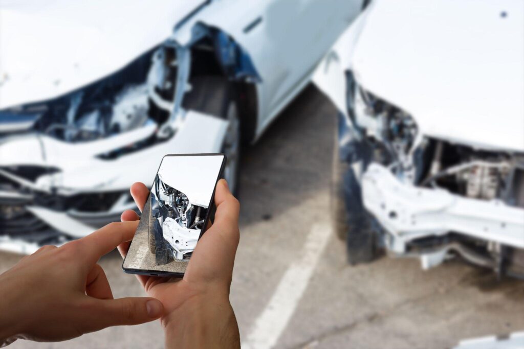 A Person Holding A Smartphone Capturing A Photo Of Two Damaged White Cars Involved In An Accident, With The Phone Screen Reflecting The Scene.