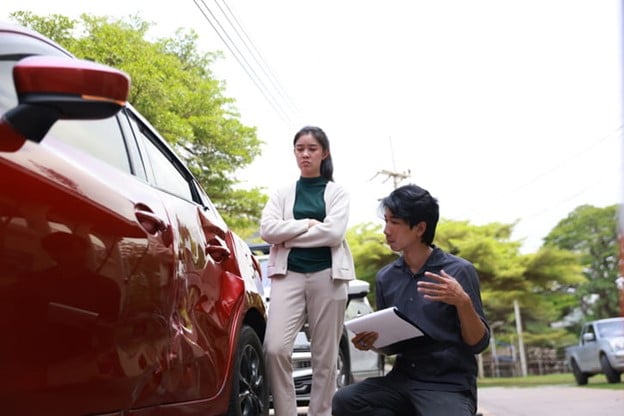 What Happens When You Get In An Accident In A Company Vehicle? 1 A Man With A Clipboard Discussing With A Woman Standing With Arms Crossed Beside A Damaged Red Company Car On A Road With Greenery In The Background.
