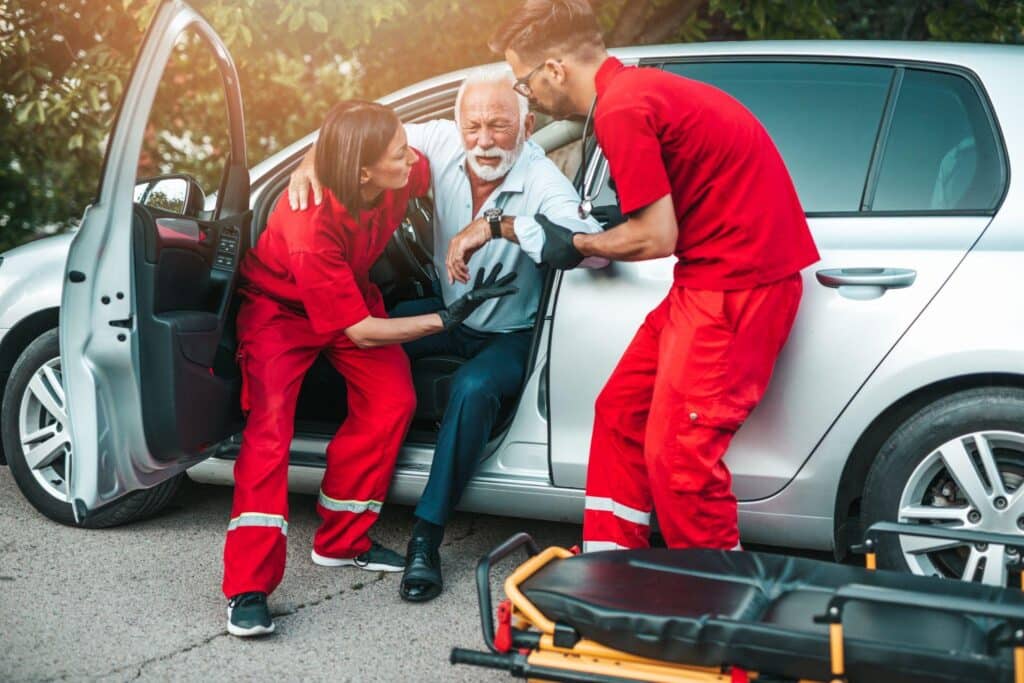 Two Paramedics In Red Uniforms Assisting An Elderly Man Inside A Silver Car With An Open Door, With A Stretcher Nearby.