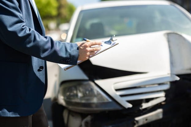 A Person In A Blue Suit Jacket Writing On A Clipboard While Inspecting A Damaged White Car With A Crumpled Front End On A Road.