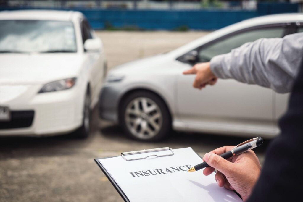 A Hand Holding A Pen Signing An Insurance Form With Two White Cars Involved In An Accident In The Background.