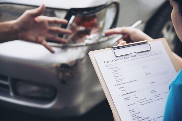 A Hand Holding A Clipboard With A Personal Accident Claim Form Being Filled Out, With A Damaged Car In The Background.