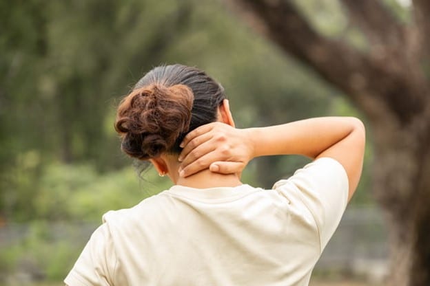 A Woman With A Bun Hairstyle Wearing A Beige Shirt, Massaging Their Neck Outdoors With A Blurred Green Background.