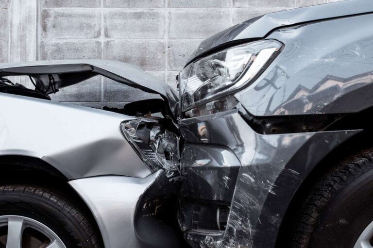 Two Damaged Cars, A Silver One And A Black One, Collided Head-On With Crumpled Fronts, Positioned Against A Concrete Wall Background.